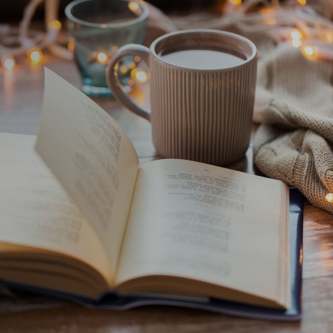 Open book on a table with a mug of tea. In the background, a tea light candle, a throw blanket and small string lights.