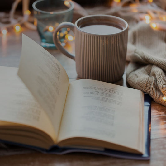 Open book on a table with a mug of tea. In the background, a tea light candle, a throw blanket and small string lights.