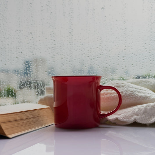 An open book and red mug with a white throw blanket on a white table. The table is against a window, covered in rain drops.