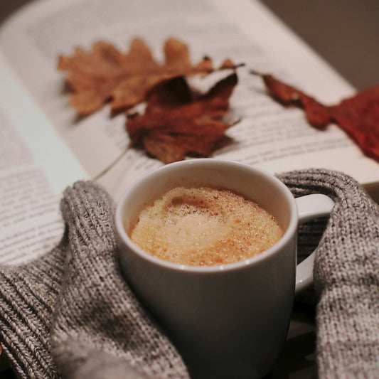 An open book with fall leaves. Hands tucked into sleeves holding a hot mug of tea with froth on top.