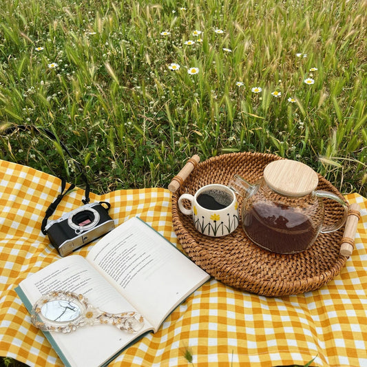 Outdoor picnic scene with a yellow checkered blanket on grass with daisies, featuring an open book, a mug of tea, a clear glass teapot, and a vintage camera on a wicker tray.