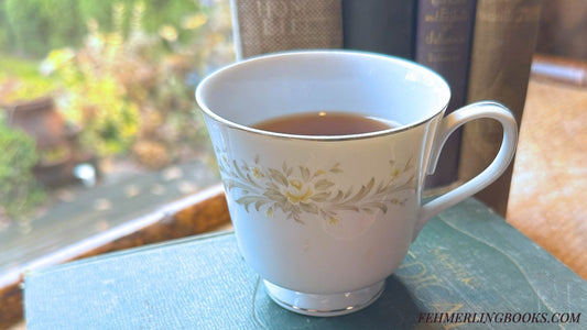 Vintage tea cup with floral print sitting with several vintage, hardcover books. Window in the backround showing a rainy day and green foliage. "FEHMERLINGBOOKS.COM" is in the lower right corner.