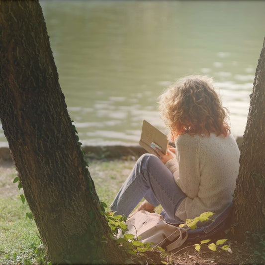 Person with curly hair sitting outdoors, leaning against a tree by a body of water, reading an open book.