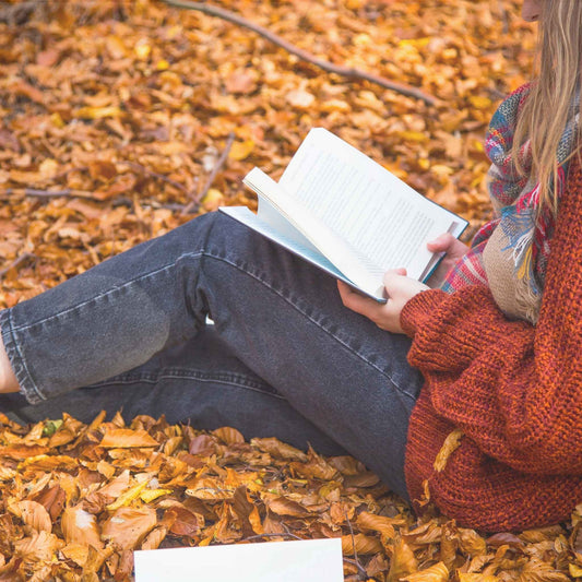 Woman in jeans and red knit sweater sitting on the ground, covered with with fallen leaves, reading a book.