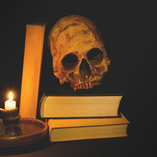 Dark black background with a stack of books with a skull resting on top, illuminated by a candle stick.