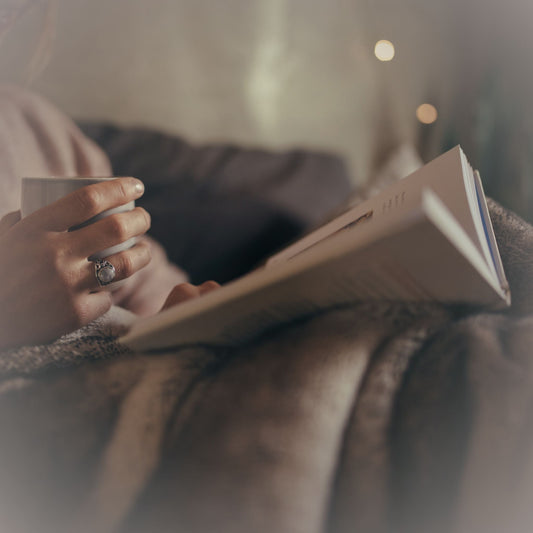 Person sitting with a cozy blanket with a cup of tea and book in their lap.