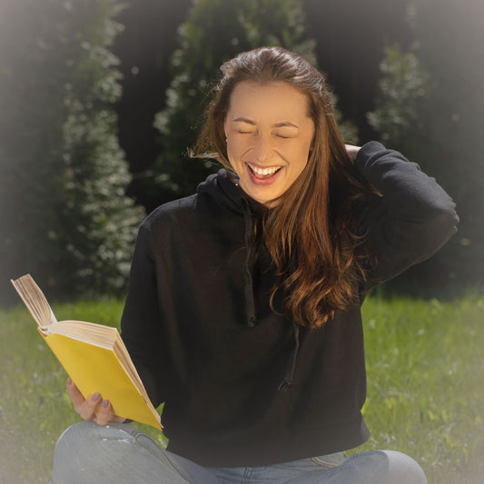Woman in a hoodie and jeans sitting cross legged on grass, laughing with a book in her hand.