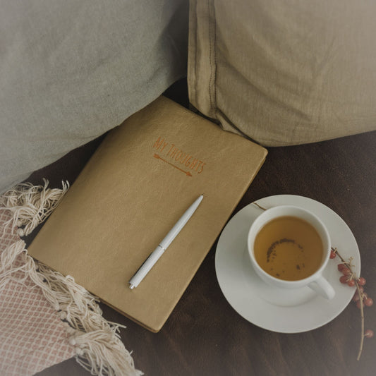 A journal with a pen on a cozy couch with a cup of tea on a saucer next to a throw blanket.