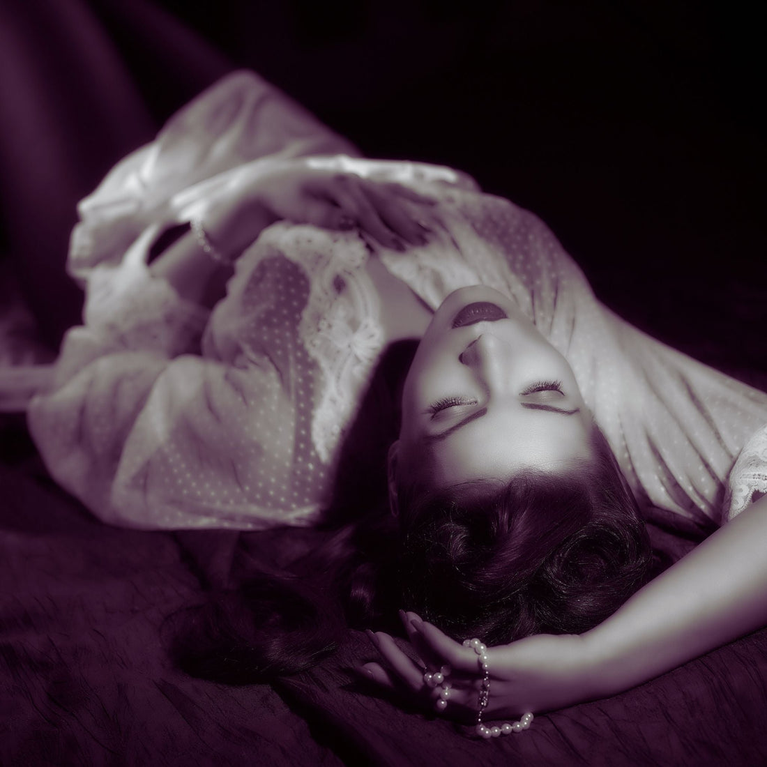 Sepia photo of woman laying on back in vintage nightgown with pearl in hand.