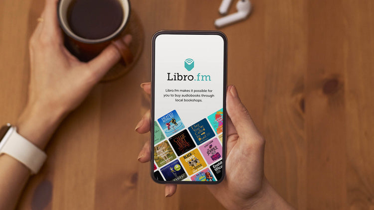 Person with a mug of tea while holding their phone with the libro.fm app open. Background is a wood table and has earbuds laying near the mug.
