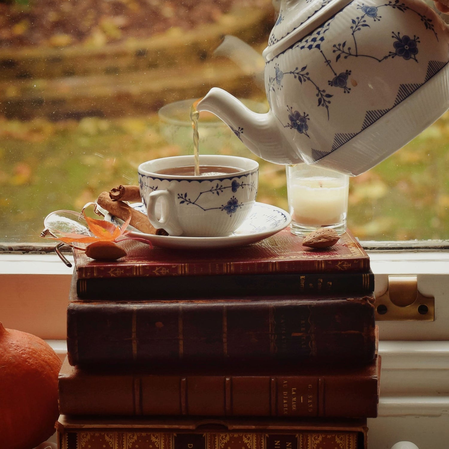 A white ceramic teapot with a blue floral pattern pours tea into a matching white teacup with a saucer, set on a stack of vintage books beside a candle and an orange, with a blurred autumn window background.