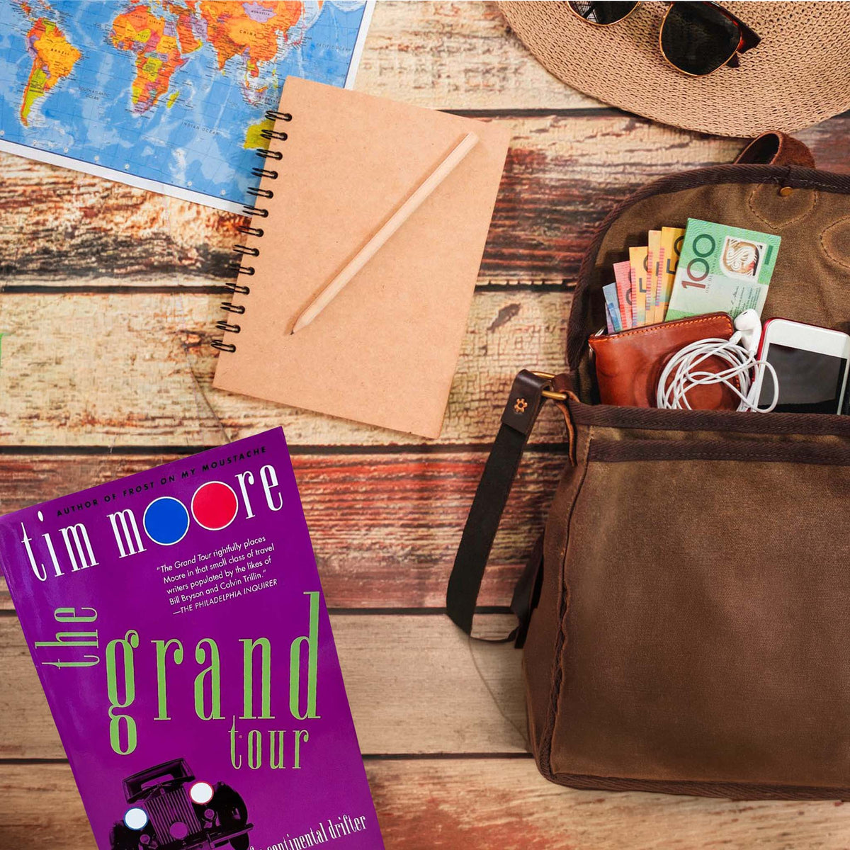 Travel flat lay on rustic wood featuring Tim Moore's book The Grand Tour, a world map, kraft notebook, straw sun hat, and a canvas backpack holding an open wallet with currency, wired earbuds, a smartphone, and a silver camera.