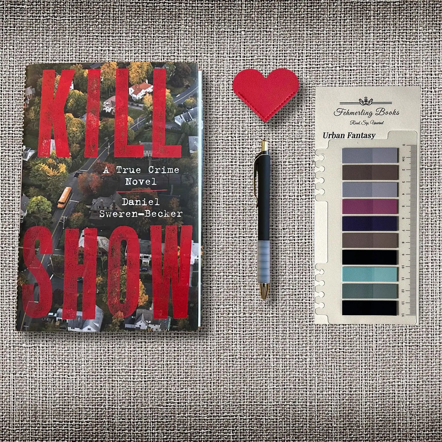 Flatlay image of a hardcover book, a pen, a heart shaped leather bookmark, and annotation tabs on a grey linen background