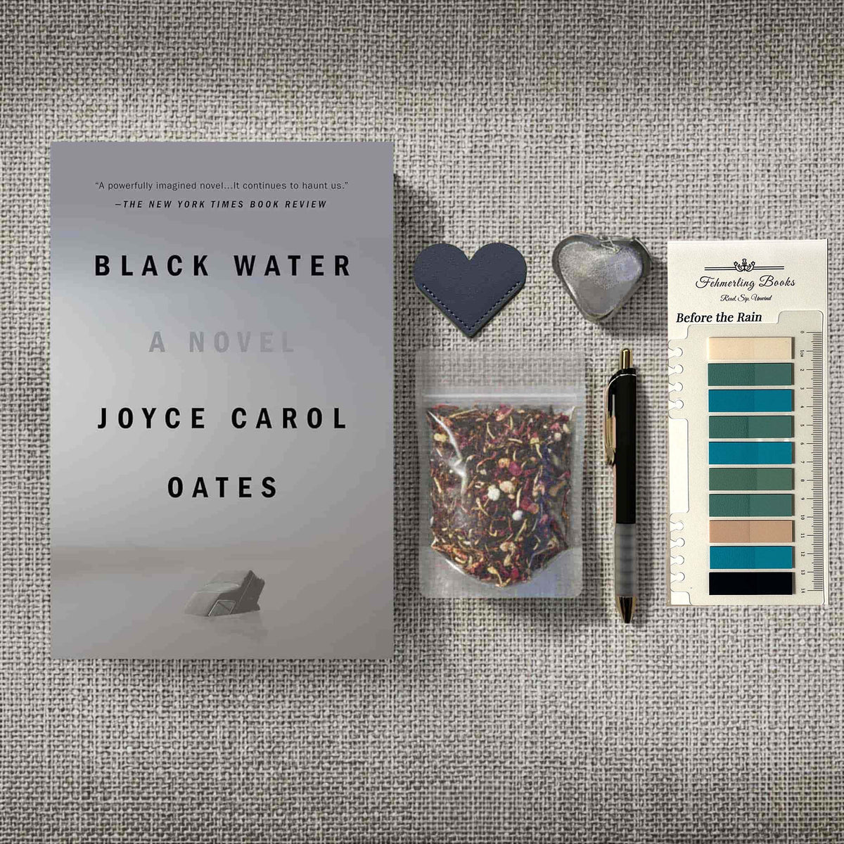 A styled flat-lay featuring the book 'Black Water' by Joyce Carol Oates alongside a small clear bag of tea, a black pen, a dark heart-shaped bookmark, a silver heart-shaped tea infuser, and annotation tabs on a textured gray fabric background.