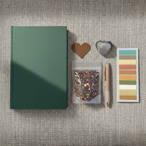 Flatlay image of a book, heart shaped leather bookmark, a pouch of loose leaf tea, a tea infuser, a pen, and annotation tabs on a linen background.