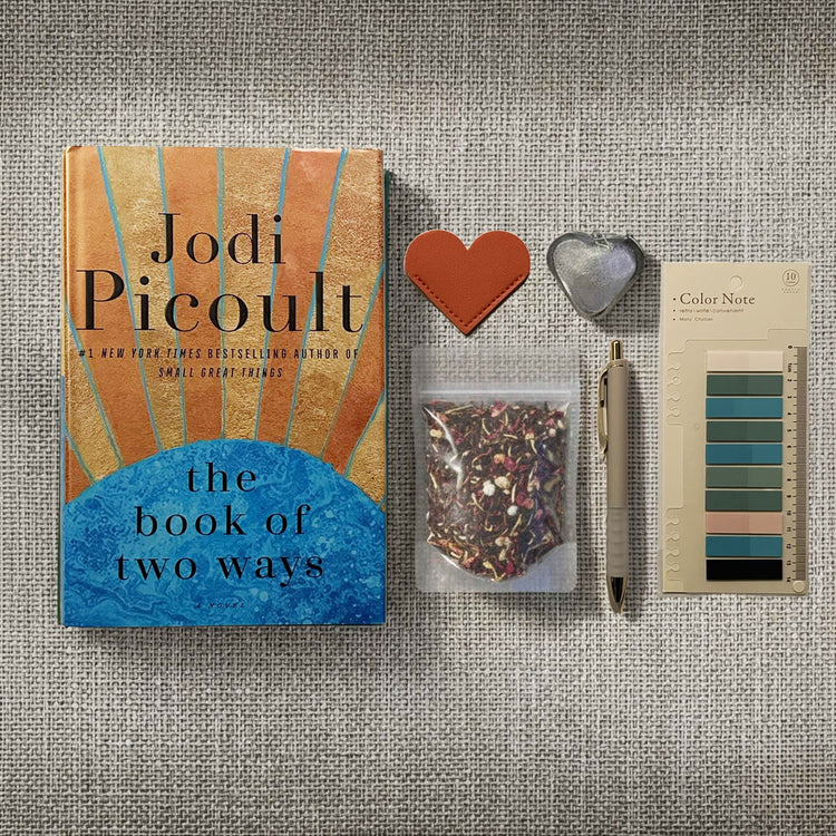 Flatlay image of a book, heart shaped leather bookmark, a pouch of loose leaf tea, a tea infuser, a pen, and annotation tabs on a linen background.
