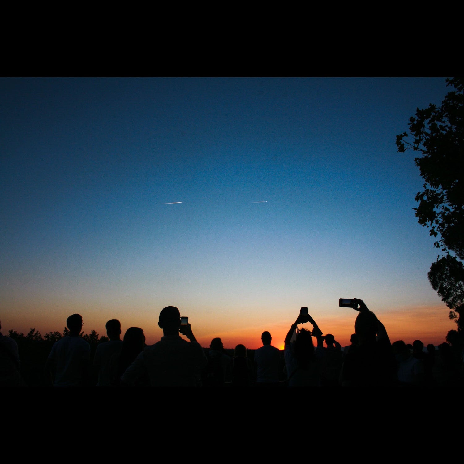Photo of people silhouetted as they use their phones to take a picture of the beautiful sunset and shooting stars in the sky.
