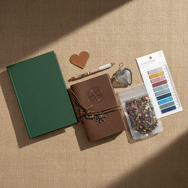Flatlay image of a book, journal, heart shaped leather bookmark, a pen, a heart shaped tea infuser, loose leaf tea, and annotation tabs on a linen background.