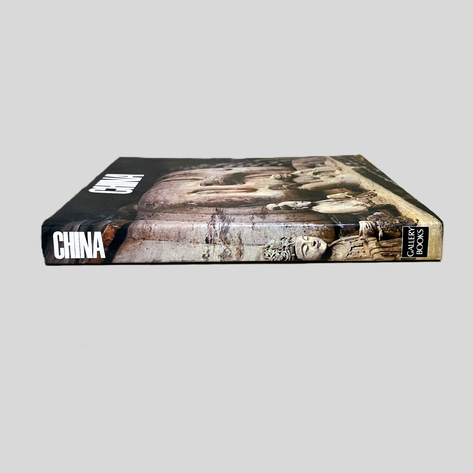 Side-angle view of the oversized volume 'China' resting on a grey surface. The screen captures the bold title and Gallery Books branding on the spine, reflecting the high-quality production of the late 20th century. The layout emphasizes the artistic and cultural significance of the Buddhist imagery featured on the dust jacket.