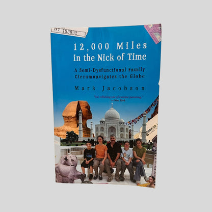 12,000 Miles in the Nick of Time by Mark Jacobson book cover. Features a vibrant blue background with a globe illustration and colorful travel-themed icons. The title and author's name are printed in bold white and yellow serif typography.