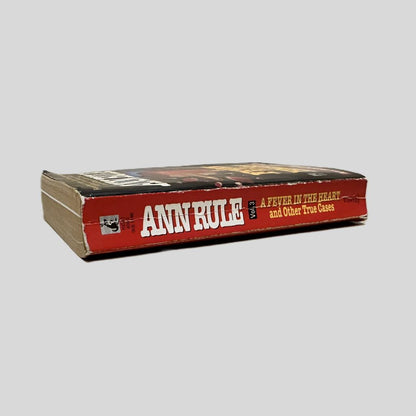 Close-up spine view of A Fever In The Heart by Ann Rule centered on a neutral light gray background. Spine shows cocking with minimal creasing.