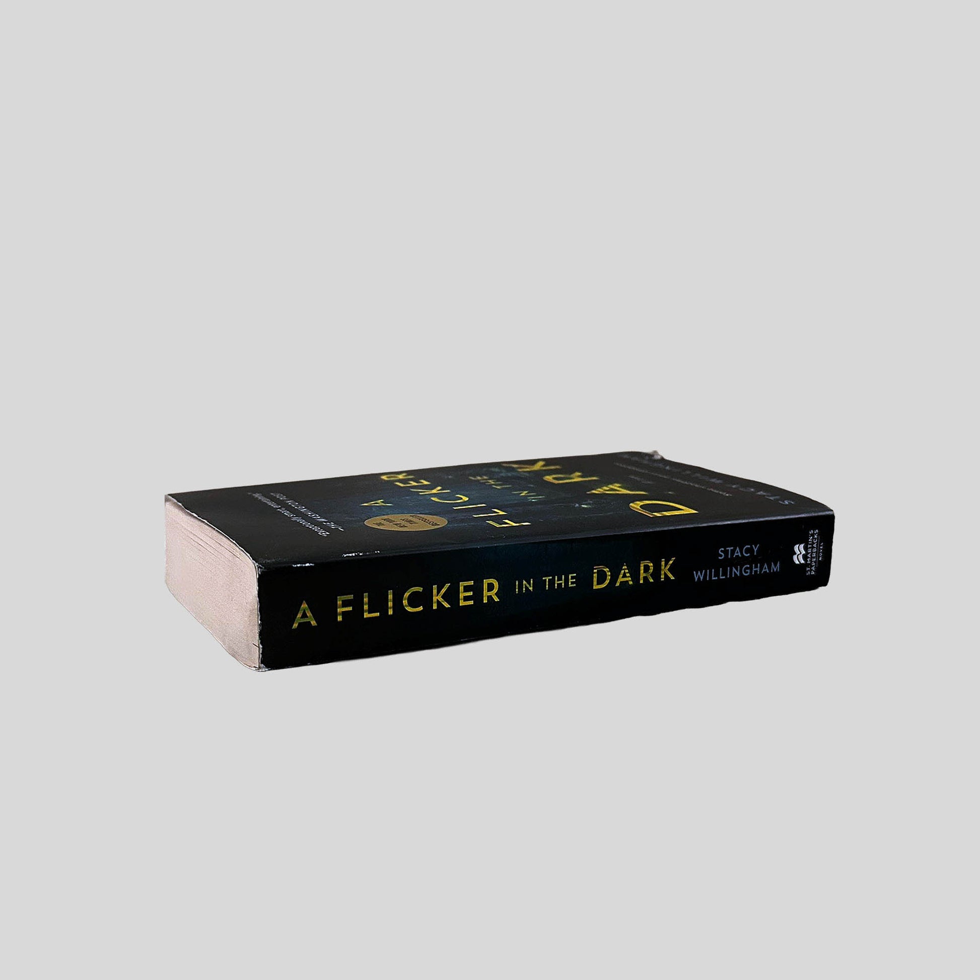 Close-up spine view of A Flicker in the Dark by Stacy Willingham centered on a neutral light gray background. Spine shows minimal creasing and chipping at the top of the spine.