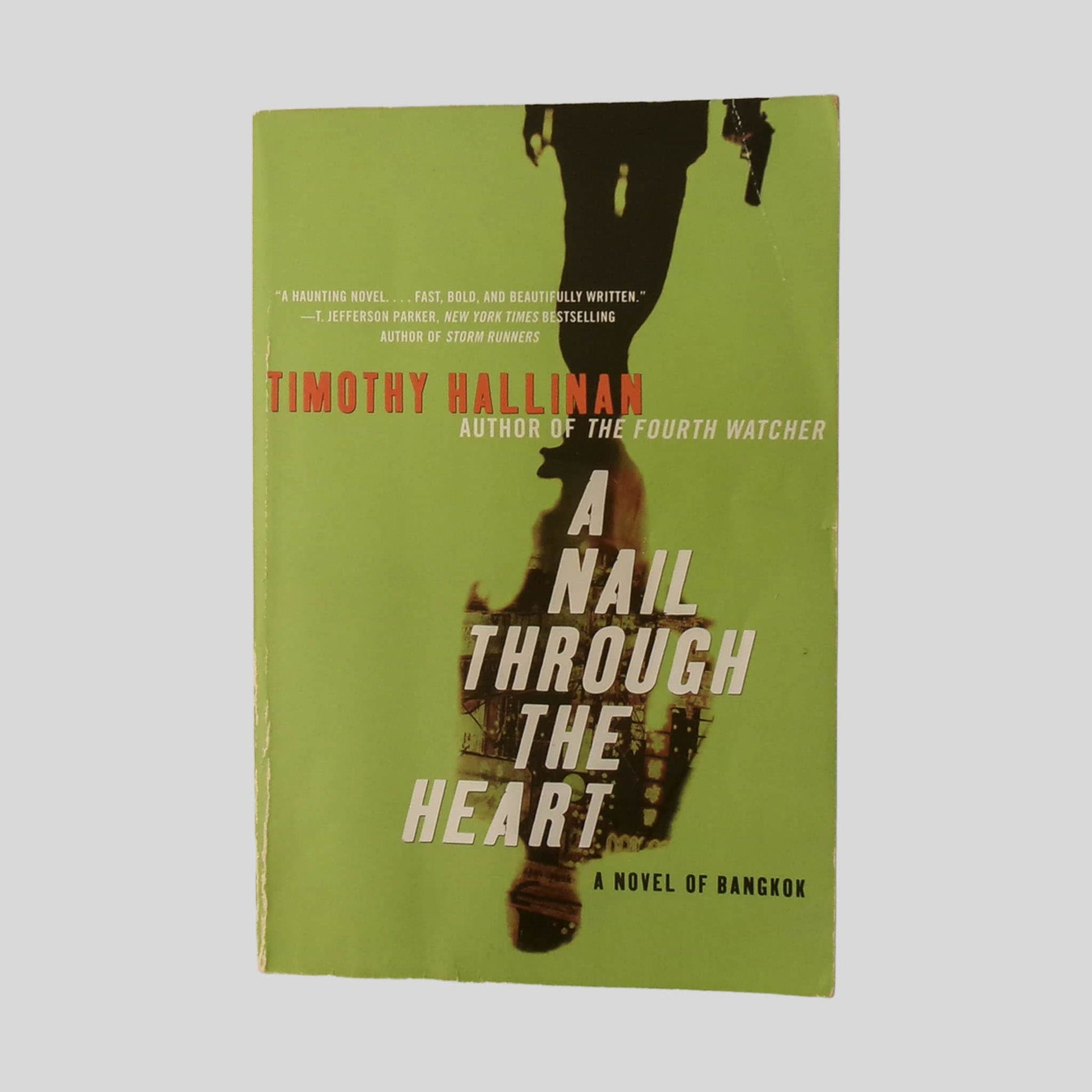 Front cover view of A Nail Through The Heart by Timothy Hallinan centered on a neutral light gray background. Chipping is present around the edges of the book. The book has a slight wave. All pages are clean. 