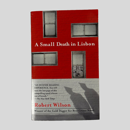Front cover of the book "A Small Death in Lisbon" by Robert Wilson, featuring a red brick building with four windows and the shadows of two figures wearing hats cast on the white wall below.