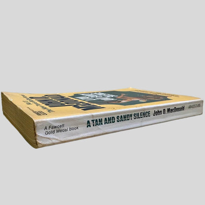 Close-up spine view of A Tan and Sandy Silence by John D. MacDonald centered on a neutral light gray background. Spine shows creasing. Pages lay flat.