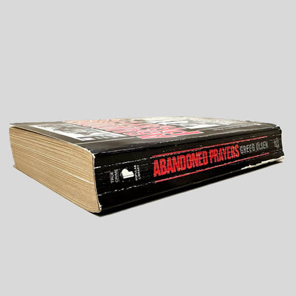 Close-up spine view of Abandoned Prayers by Gregg Olsen centered on a neutral light gray background. Spine shows creasing and chipping.