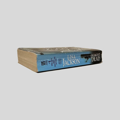 Close-up spine view of Almost Dead by Lisa Jackson centered on a neutral light gray background. Spine shows creasing. Pages lay flat.
