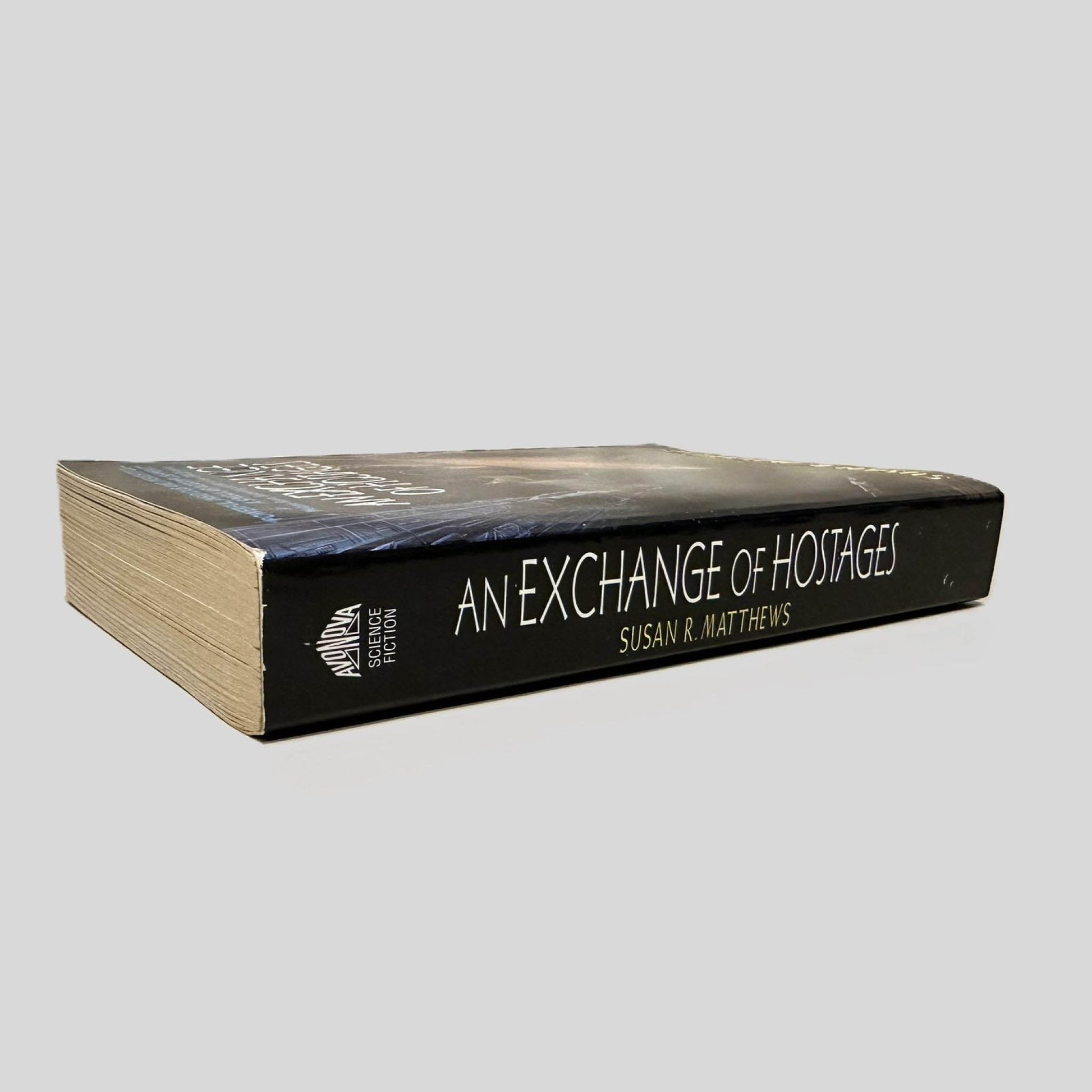 Close-up spine view of An Exchange of Hostages by Susan R. Matthews centered on a neutral light gray background. Spine is square and shows no creasing. Pages lay flat.