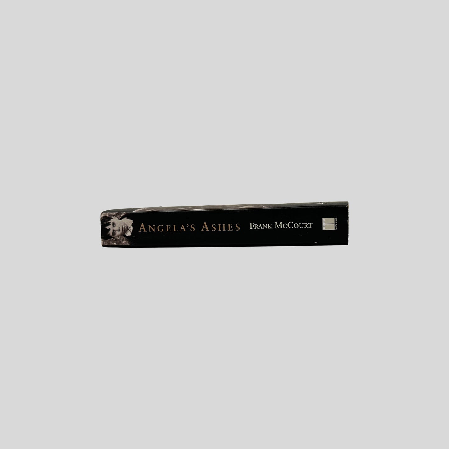 The black spine of the paperback book "Angela's Ashes" by Frank McCourt, featuring gold title text and a small portrait of a young boy's face.