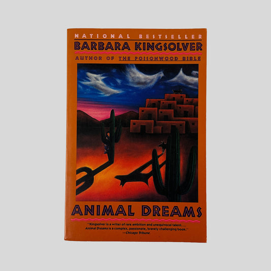 The orange front cover of the paperback Animal Dreams by Barbara Kingsolver, featuring a southwestern-style illustration of saguaro cacti, adobe buildings, and a sunset under a swirling blue sky.