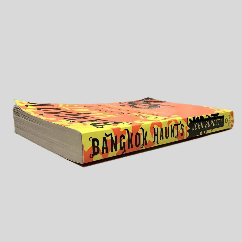 Close-up spine view of Bangkok Haunts by John Burdett centered on a neutral light gray background. Spine shows no creasing. Pages have creasing in the top 50 pages, some water damage to pages causing some waves but text is unaffected.