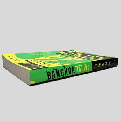 Close-up spine view of Bangkok Tattoo by John Burdett centered on a neutral light gray background. Spine shows no creasing. Some water damage to pages causing some waves but text is unaffected.