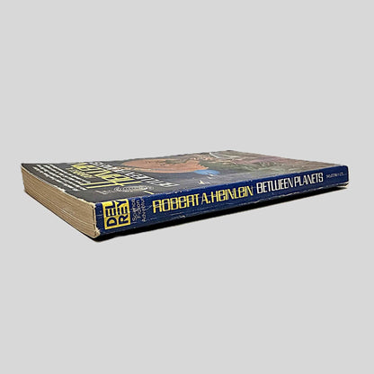 Close-up spine view of Between Planets by Robert A. Heinlein centered on a neutral light gray background. Spine shows creasing; chipping at the top and lower end of spine. Pages lay flat.