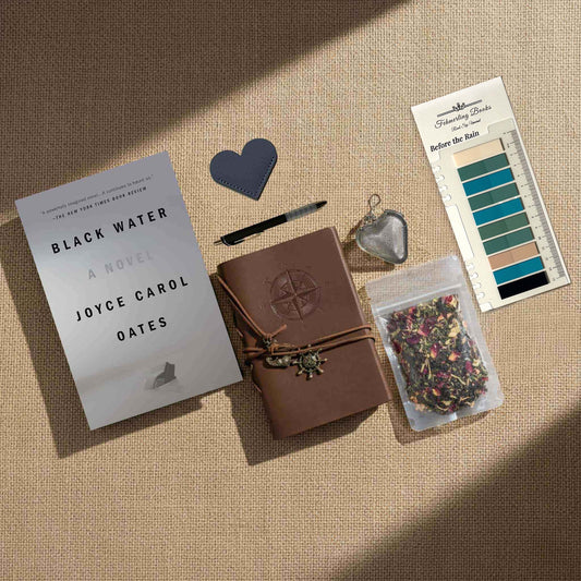 Top-down view of a curated reading set for the novel Black Water by Joyce Carol Oates, arranged on a textured beige background. The bundle includes a leather-bound journal, loose-leaf tea, a heart infuser, a pen, a bookmark, and a "Before the Rain" palette displaying translucent tabs in deep greens, teals, warm tans, and onyx black.