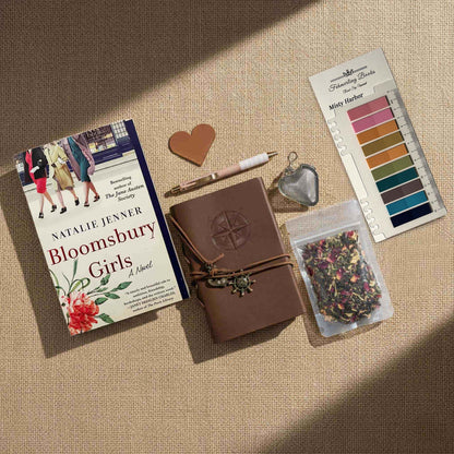 Overhead view of the "Bloomsbury Girls" Ultimate Literary Collection featuring the hardcover book by Natalie Jenner, a brown leather-bound journal with compass embossing and nautical charms, a heart-shaped tea infuser, a pouch of loose-leaf herbal tea, and a "Misty Harbor" annotation tab palette on a burlap surface.