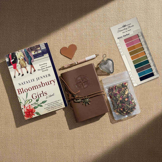 Overhead view of the "Bloomsbury Girls" Ultimate Literary Collection featuring the hardcover book by Natalie Jenner, a brown leather-bound journal with compass embossing and nautical charms, a heart-shaped tea infuser, a pouch of loose-leaf herbal tea, and a "Misty Harbor" annotation tab palette on a burlap surface.