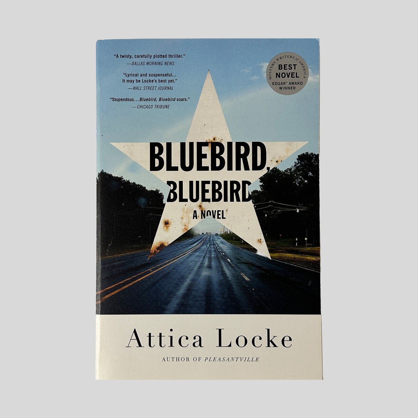 The front cover of "Bluebird, Bluebird" by Attica Locke, featuring a large, rusted white star superimposed over a photograph of a long, wet highway stretching toward the horizon.