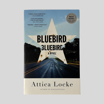 The front cover of "Bluebird, Bluebird" by Attica Locke, featuring a large, rusted white star superimposed over a photograph of a long, wet highway stretching toward the horizon.