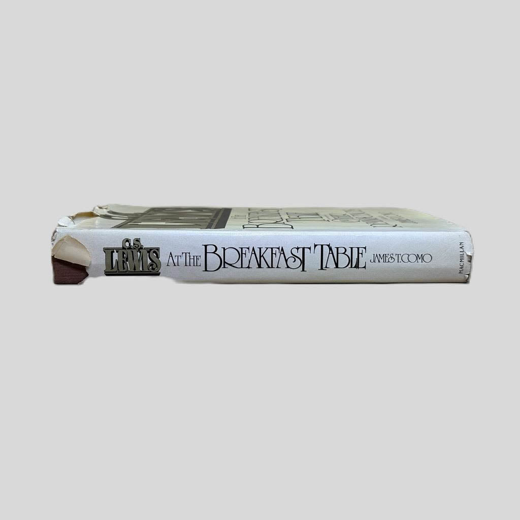Close-up spine view of C.S. Lewis At the Breakfast Table Edited by James T. Como centered on a neutral light gray background. Dust jacket is present and show significant tears at the top end of the spine; smaller tears to the jacket on the lower end. Pages lay flat.