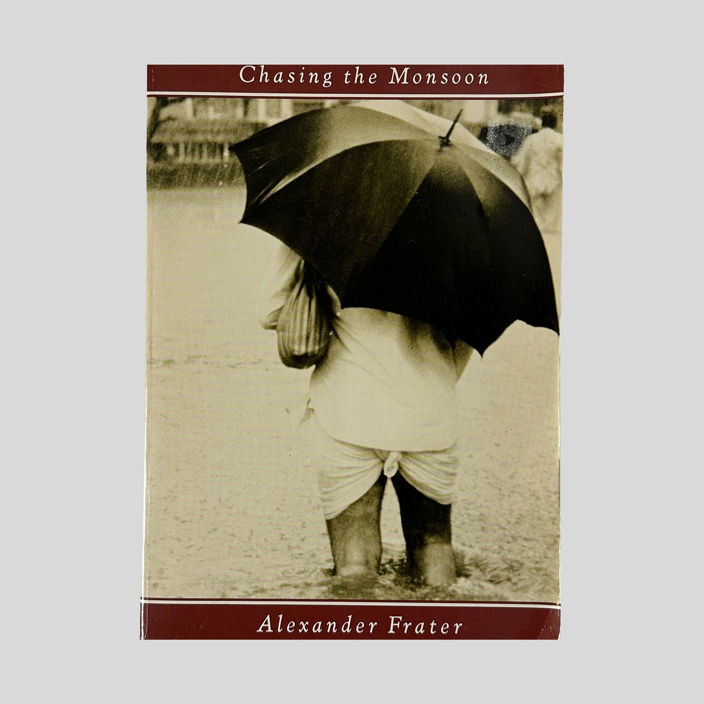 Front cover of the book "Chasing the Monsoon" featuring a sepia-toned photograph of a person in high water holding a large black umbrella, framed by maroon banners.