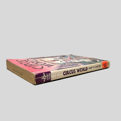 Close-up spine view of Circus World by Barry B. Longyear centered on a neutral light gray background. Spine shows creasing and rubbing. Pages lay flat. The letter "C" on a round, orange sticker is taped to the bottom of the spine for library cataloging. 