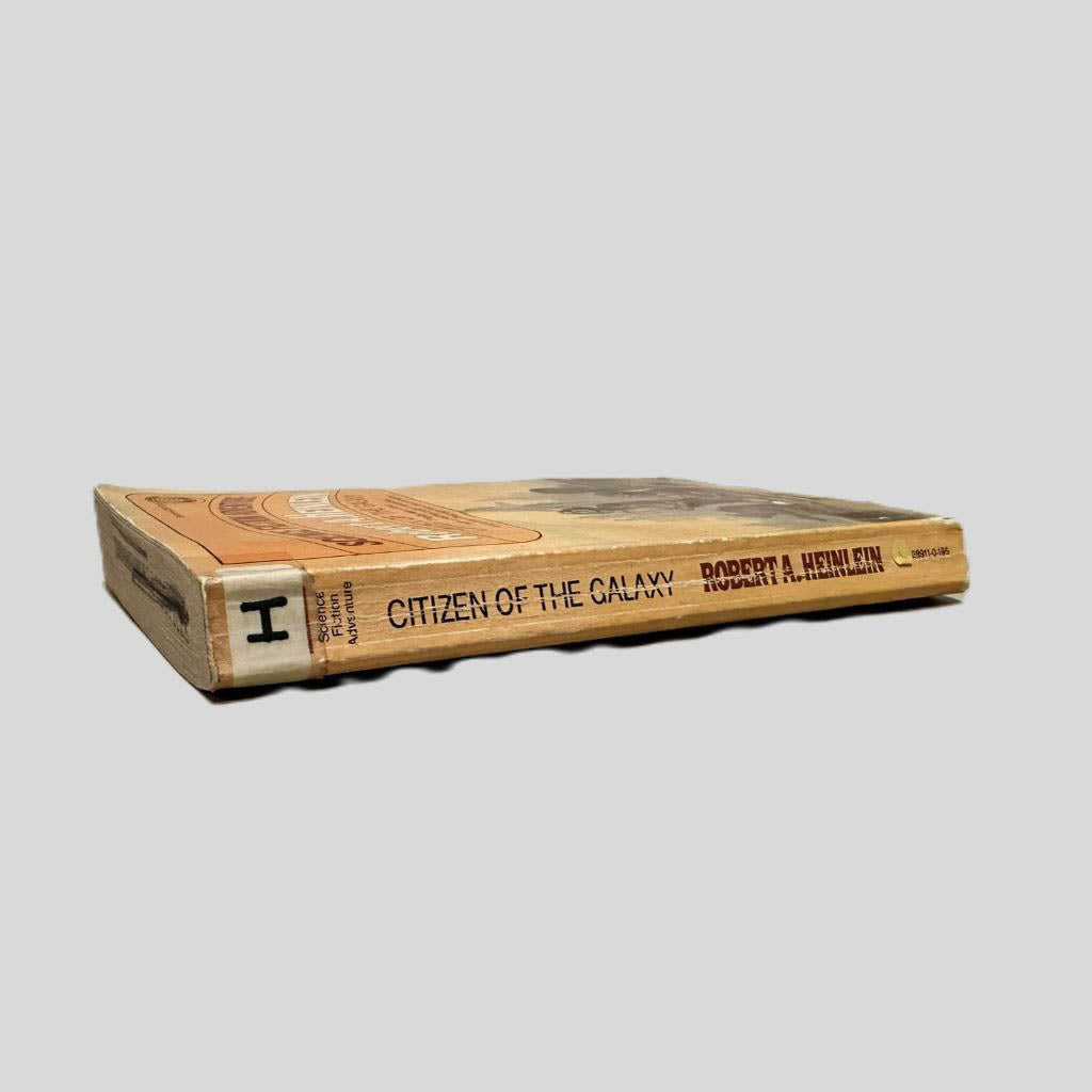 Close-up spine view of Citizen of the Galaxy by Robert A. Heinlein centered on a neutral light gray background. Spine shows creasing. Library shelving sticker is taped to the top end of the spine. Pages lay flat.