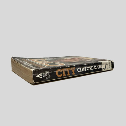 Close-up spine view of City by Clifford D. Simak centered on a neutral light gray background. Spine shows creasing and chipping. Library shelving stickers are present at the bottom of the spine. Pages lay flat.