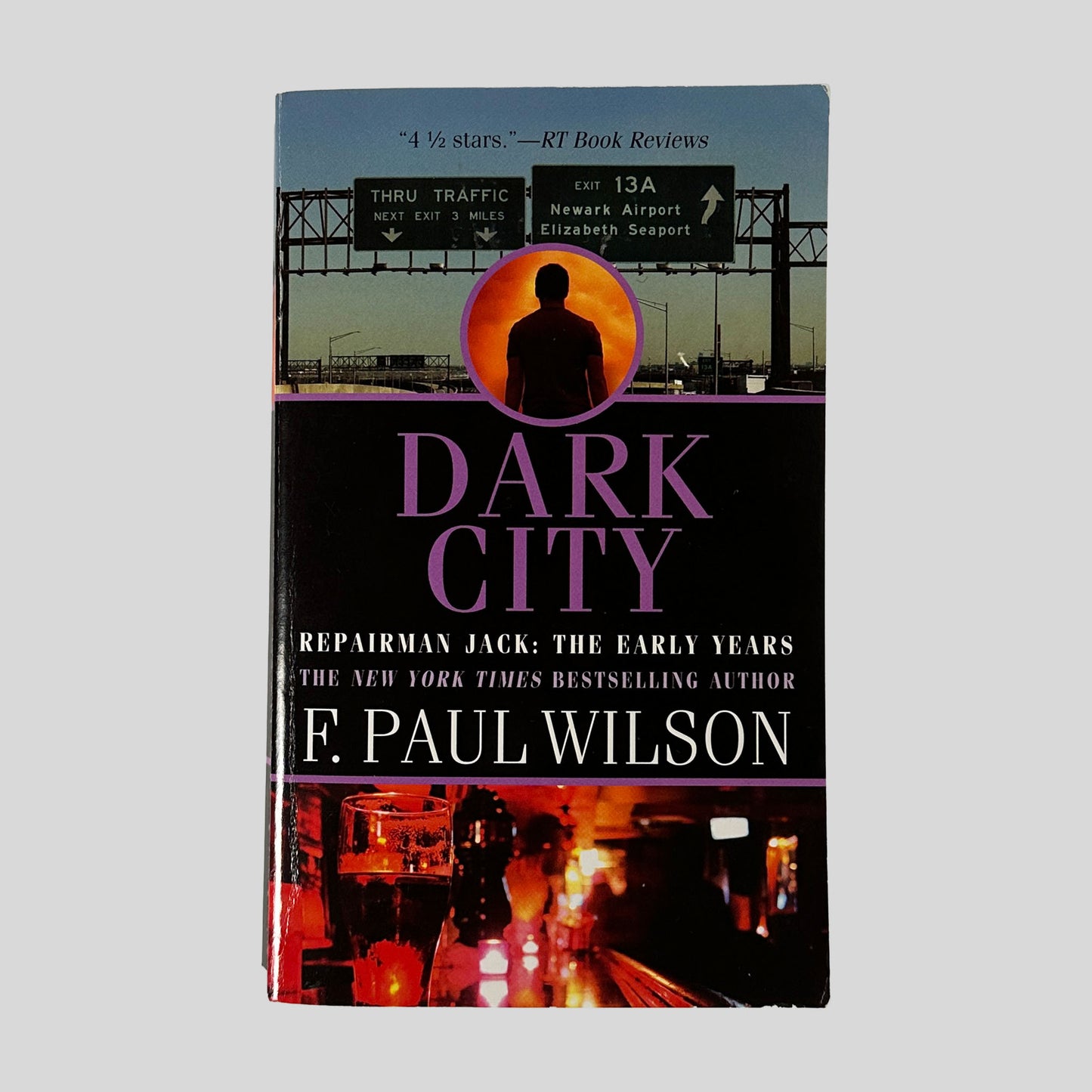 The front cover of the paperback Dark City by F. Paul Wilson, featuring a sunset-lit highway sign for Newark Airport above a photo of a pint of beer in a dark bar.