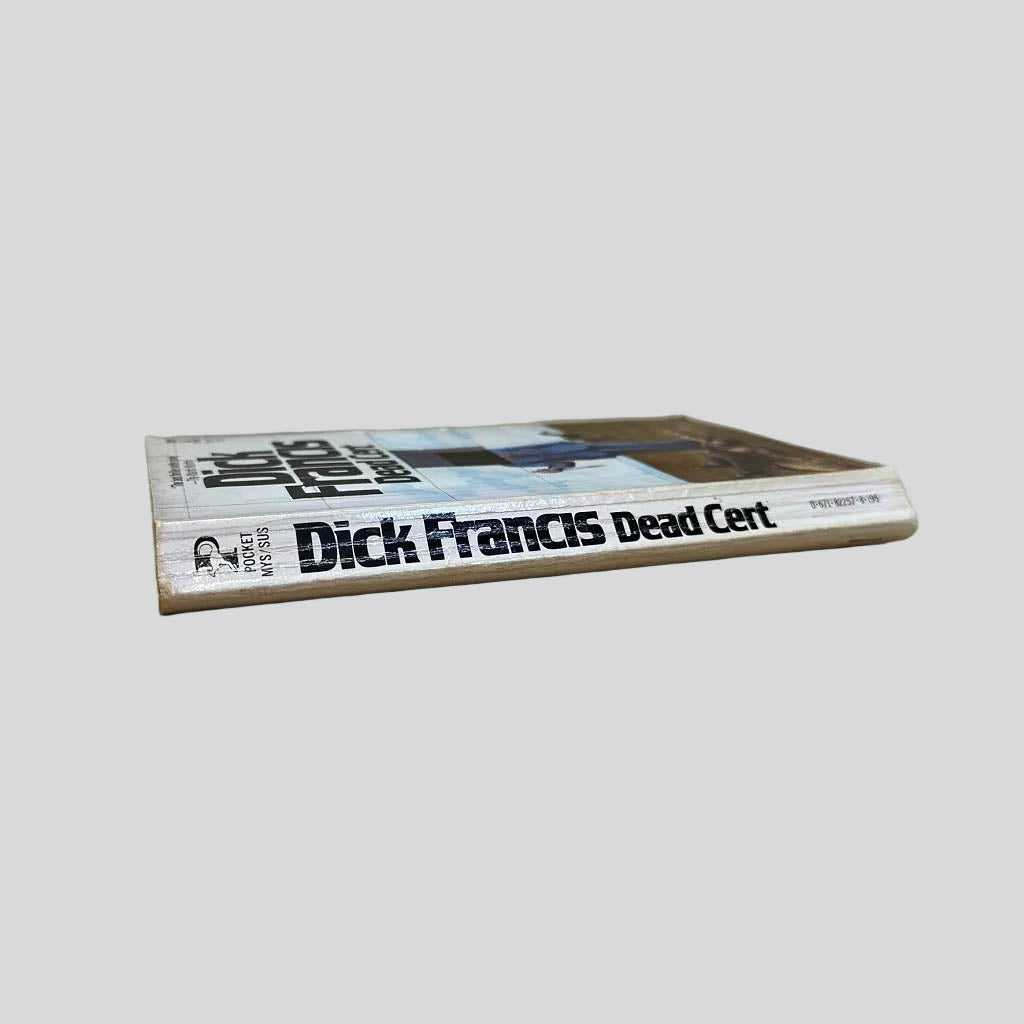 Close-up spine view of Dead Cert by Dick Francis centered on a neutral light gray background. Spine shows creasing and cocking.