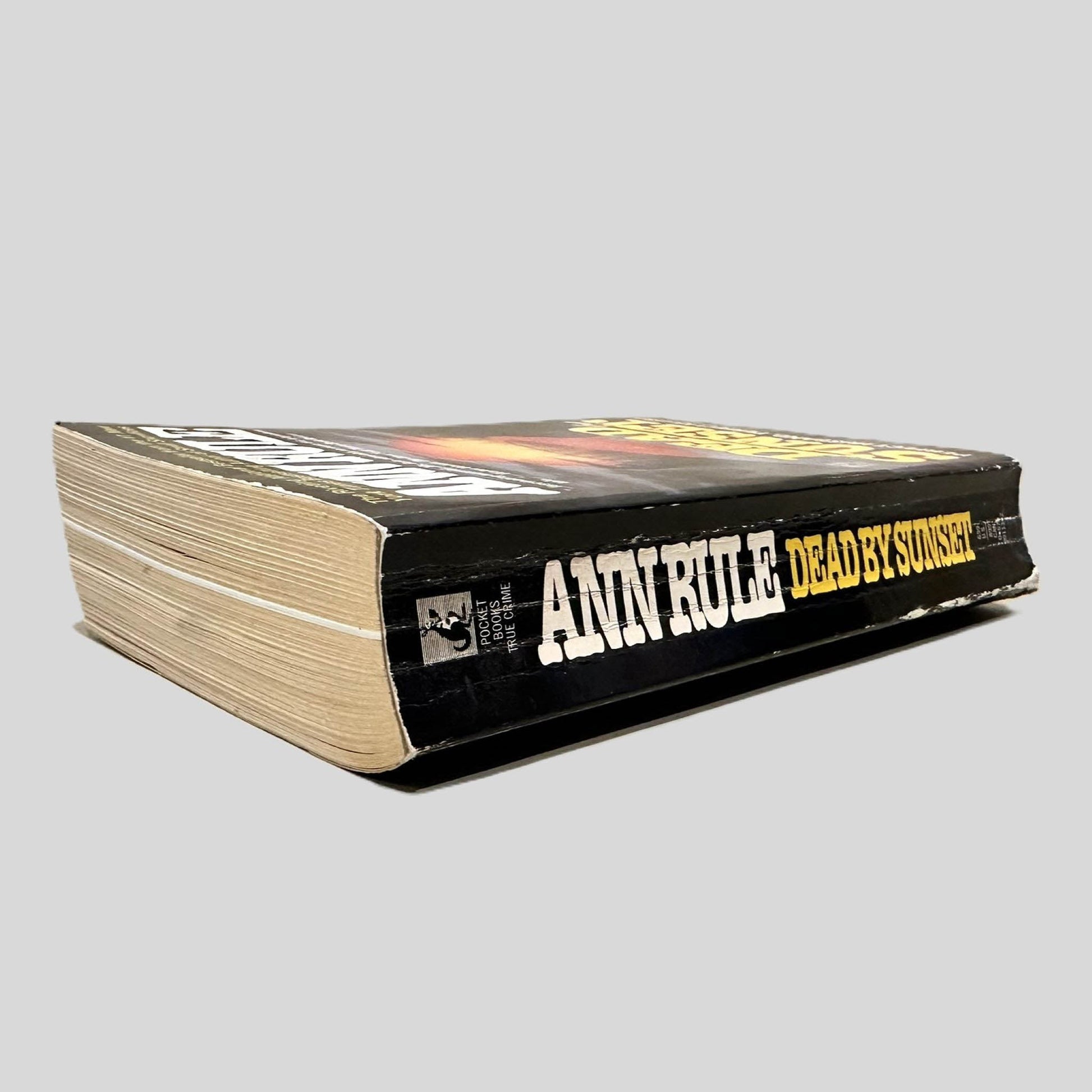 Close-up spine view of Dead by Sunset by Ann Rule centered on a neutral light gray background. Spine shows creasing.
